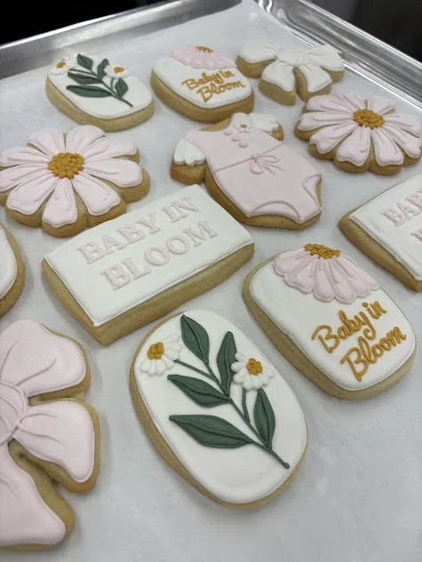 Cookies decorated with flowers, baby onesies, and the phrases "Baby in Bloom" and "Baby Bloom" in pastel pink, white, and yellow icing.