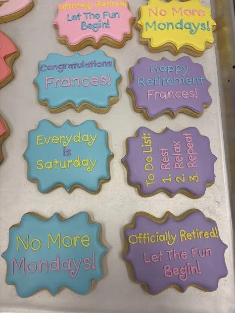 Colorful retirement and weekend-themed cookies with various phrases like "Officially Retired! Let The Fun Begin!" and "No More Mondays!" arranged on a table.