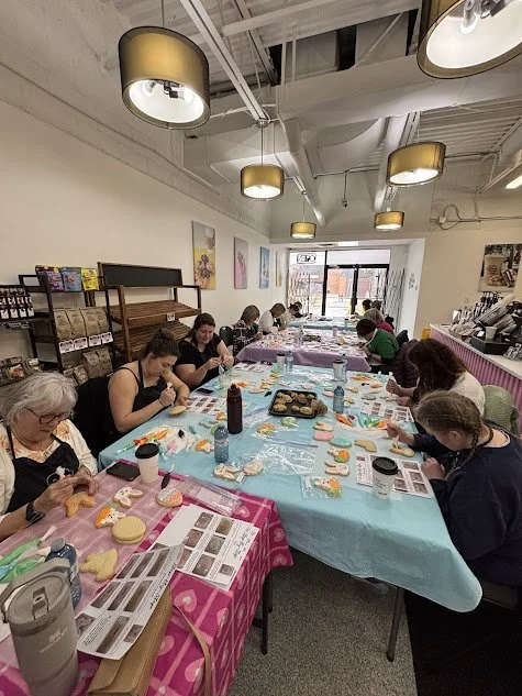 People sitting at a long table decorating cookies in a bright room with posters on the walls and large windows.