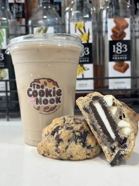 A cold coffee drink in a clear cup with a clear lid, labeled 'The Cookie Nook', placed next to two cookies, one whole and one sliced in half to show layered cookies and cream filling inside.