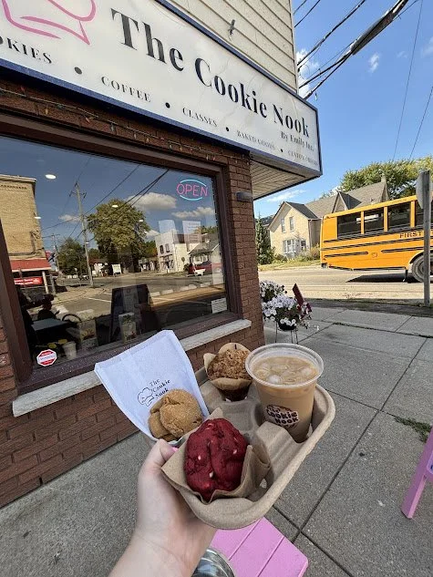 Person holding a tray with cookies, ice cream, and a coffee outside a bakery called 'The Cookie Nook' with a yellow school bus in the background.