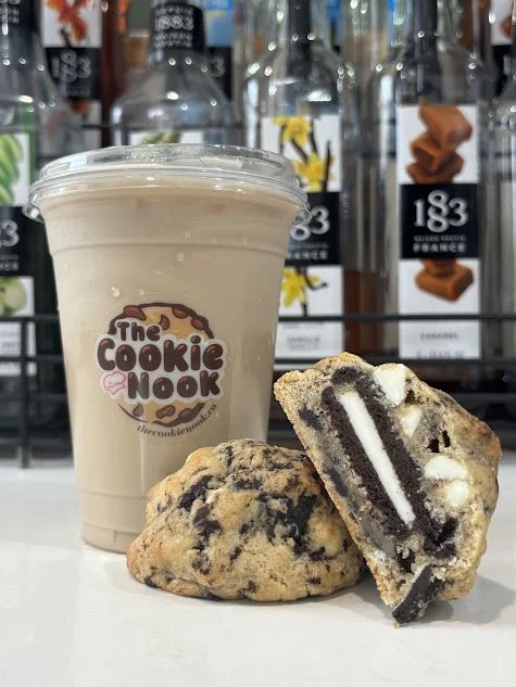 A cup of iced coffee with the logo 'The Cookie Nook' on the front, placed next to two cookies, one whole and one cut in half showing an Oreo filling inside, on a store shelf with bottled drinks in the background.