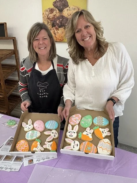 Two women smiling at a table with decorated cookies shaped like bunnies and eggs, in front of a yellow wall with a baked goods poster.
