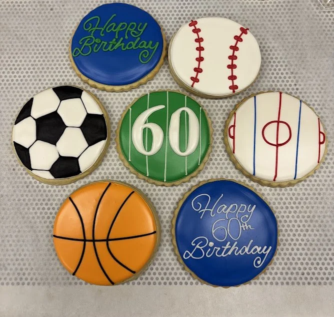 Seven decorated cookies celebrating a 60th birthday, featuring logos of a soccer ball, baseball, hockey rink, basketball, and birthday messages.