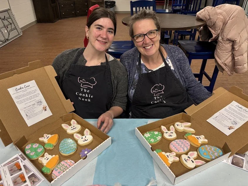 Two women sitting at a table with bunny and decorated cookies in open boxes, wearing aprons with a cookie chef logo, smiling at the camera.
