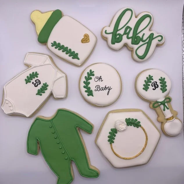 Decorative cookies with baby-themed designs in white, green, and gold, including a baby bottle, the word "baby," a onesie, a bowtie, and a pacifier.