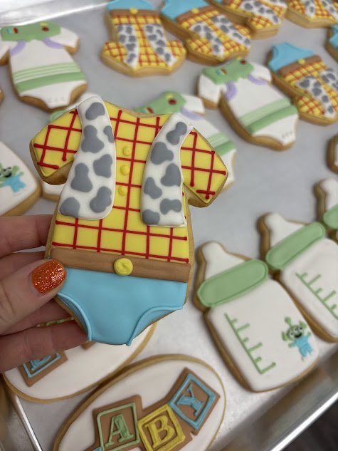 Decorative cookies shaped like baby clothes, including a yellow onesie with a red grid pattern, gray and white striped socks, and other baby-themed cookies.