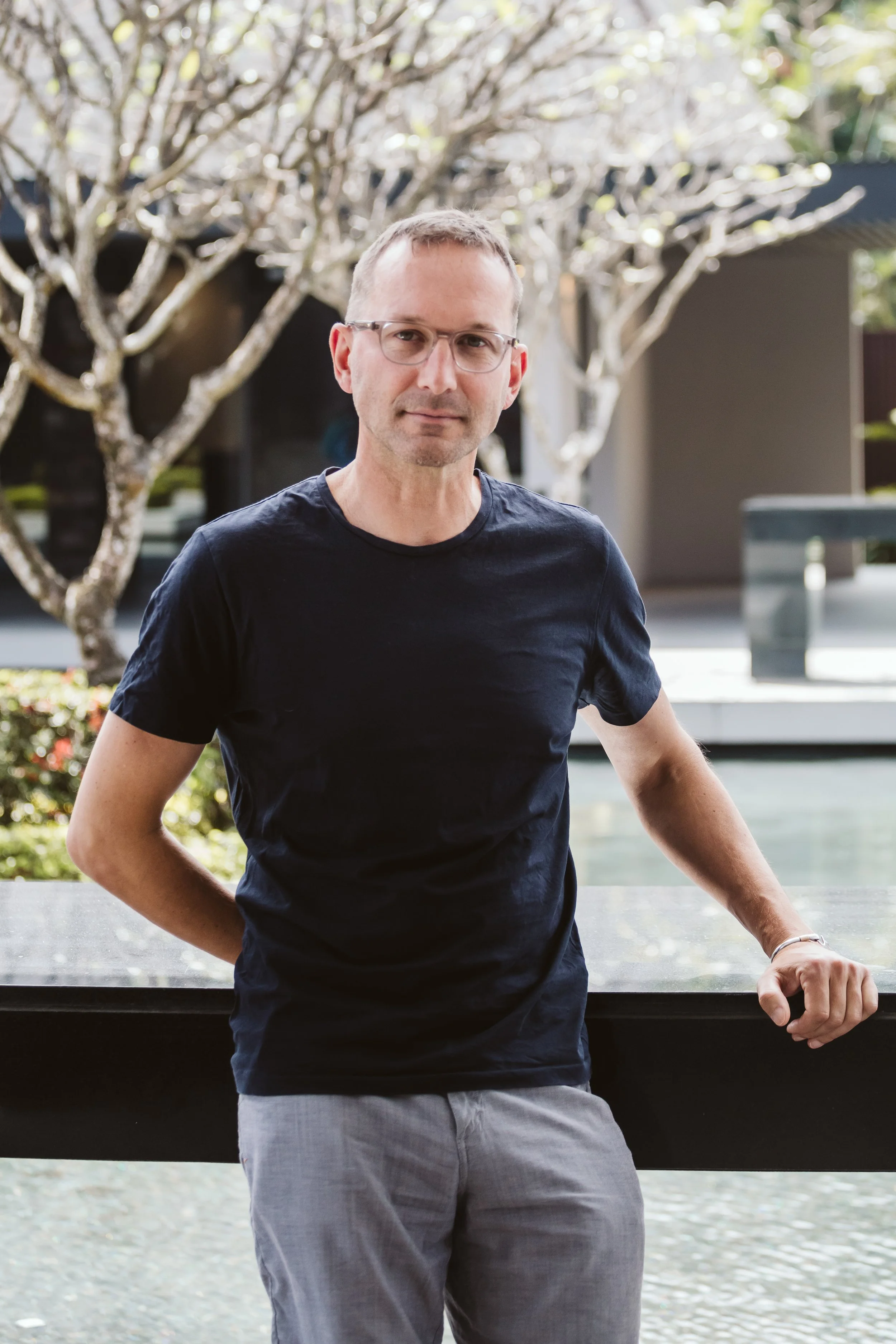 Man wearing glasses, black t-shirt, and light gray pants, standing outdoors with trees and modern buildings in the background.