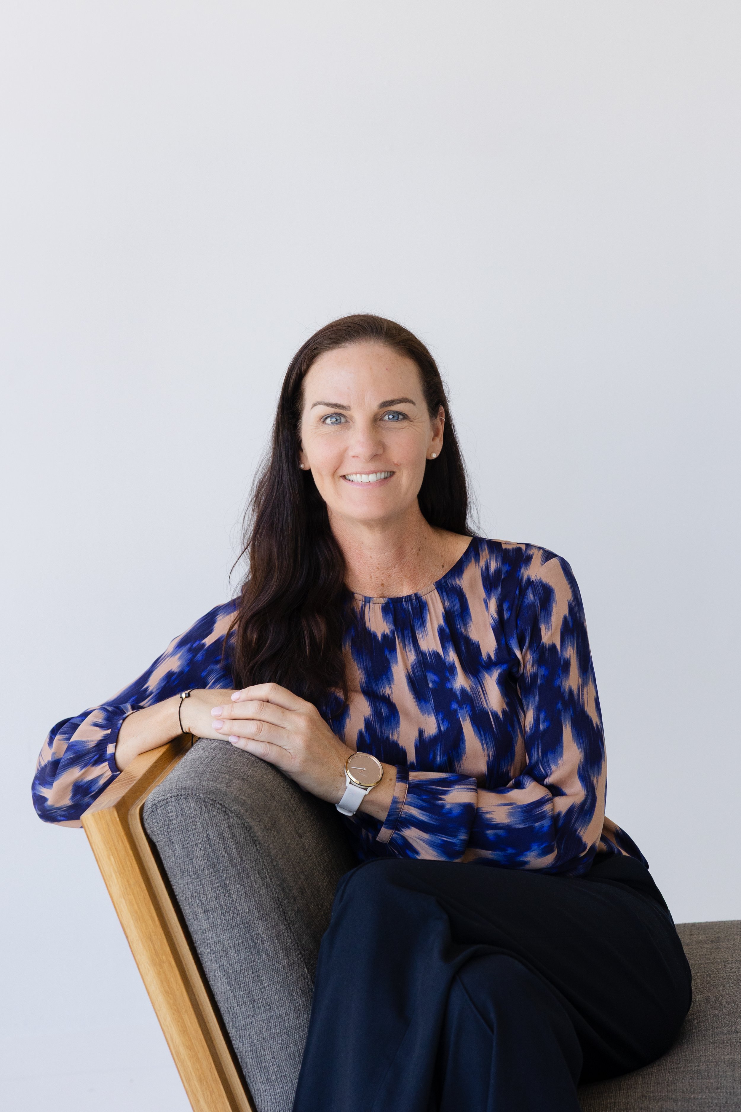 A woman with long dark hair and blue eyes, wearing a blue and beige patterned blouse, sitting on a chair with a wooden frame and gray fabric. She is smiling and has her hands resting on her lap, wearing a white watch and a bracelet against a plain light gray background.