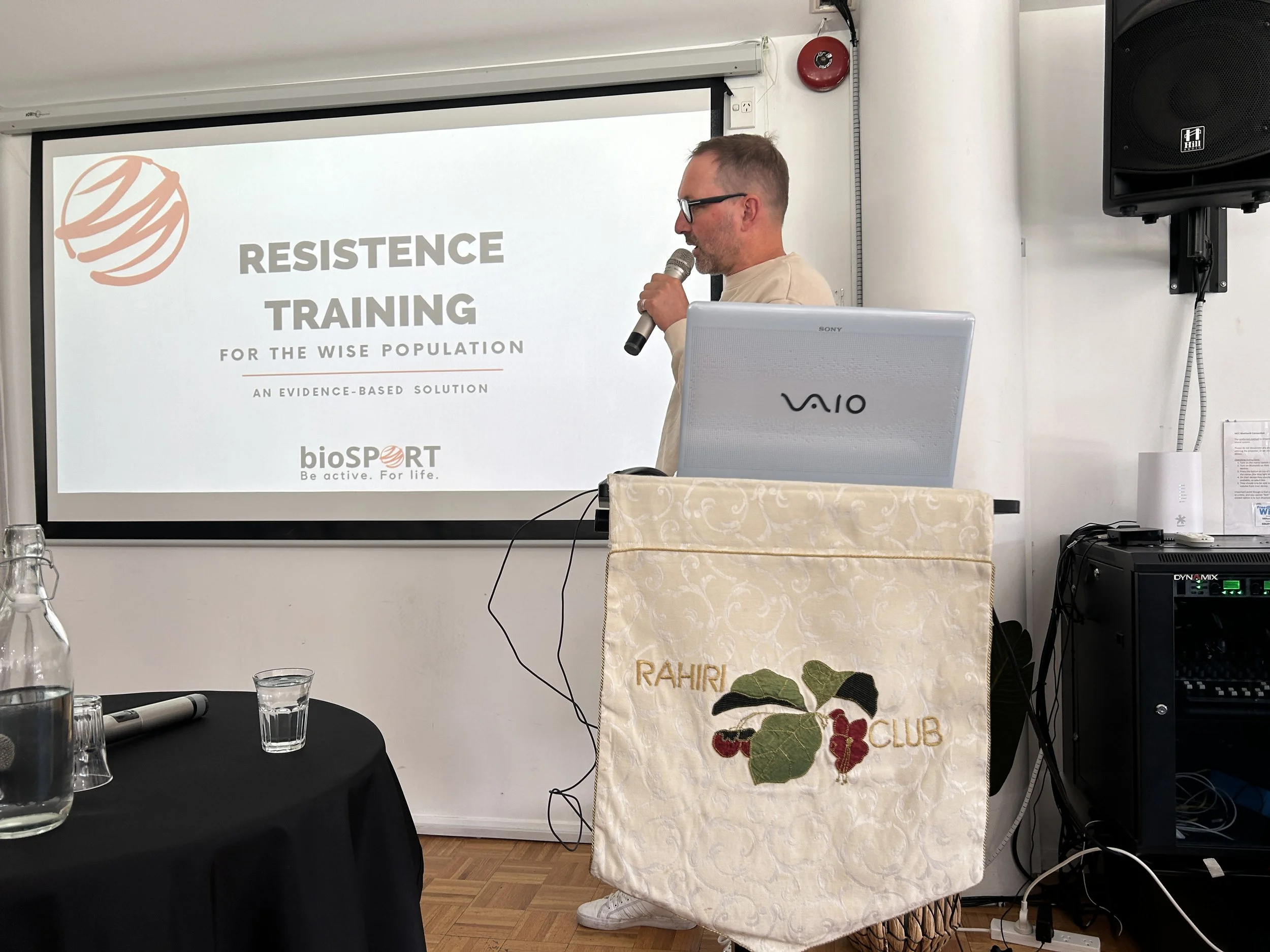 A man with glasses giving a presentation about resistance training at a conference, standing next to a projected screen displaying a logo and event information, with a laptop and a table with water glasses in the foreground.