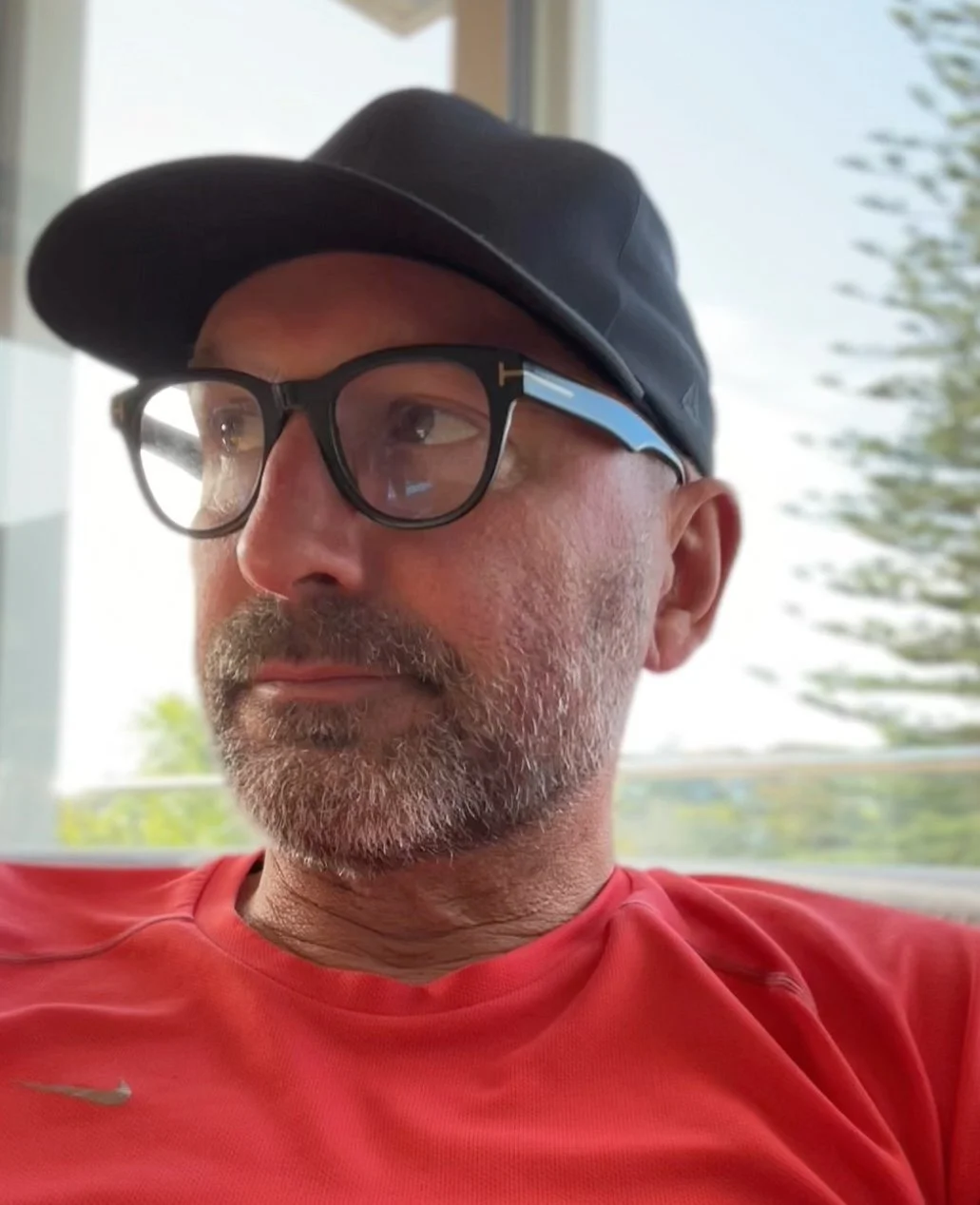 Close-up of a man wearing a black cap, black glasses, and a red shirt, sitting indoors near a window with blurred outdoor scenery.