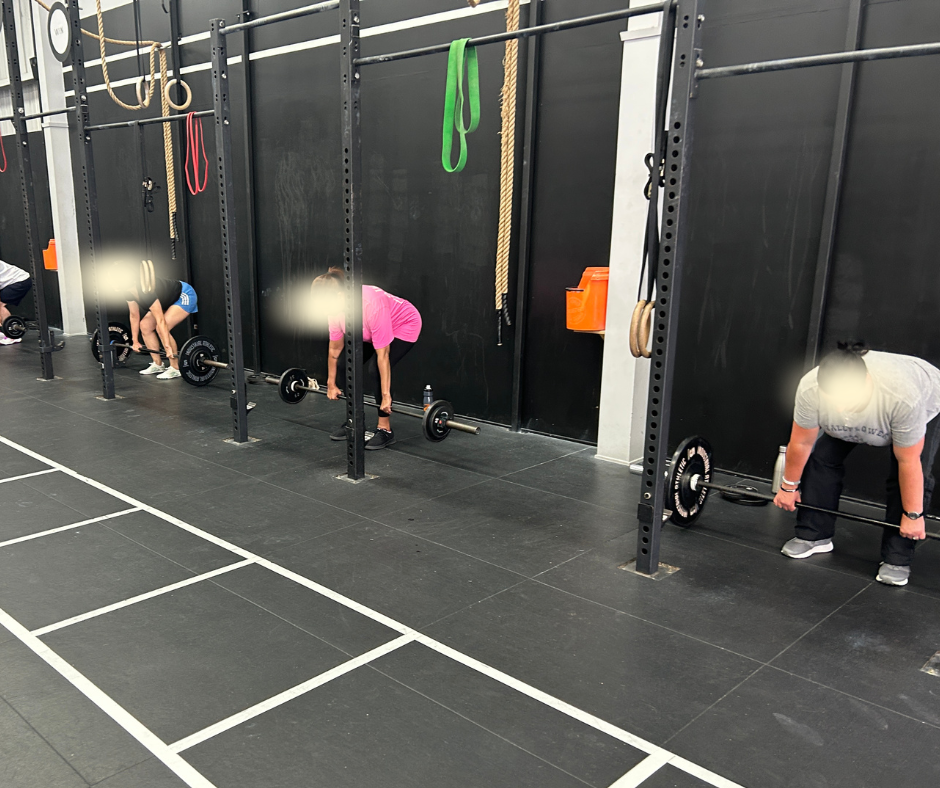 People doing deadlifts in a gym, standing in front of black wall-mounted racks with barbells, gym ropes, and resistance bands hanging.