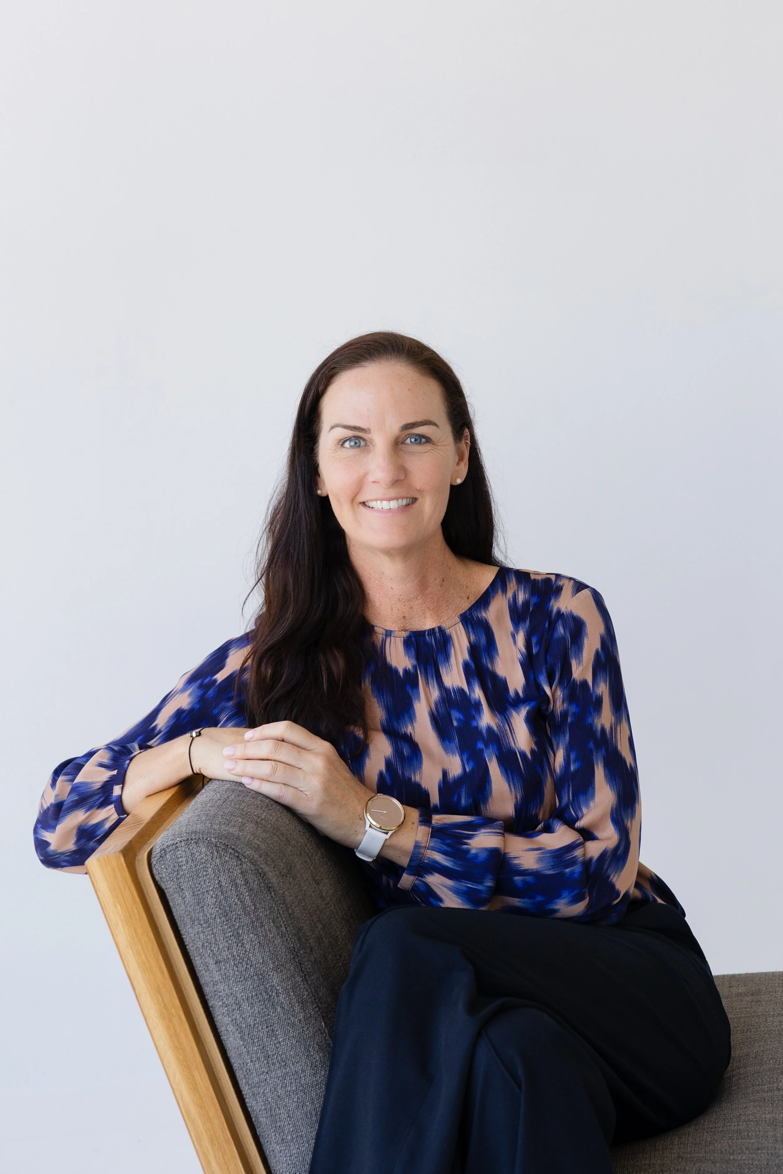 A woman with long brown hair, blue eyes, and fair skin is sitting on a mid-century modern chair with a wooden frame and gray fabric. She is wearing a blue, beige, and black patterned blouse, black pants, a white watch, and pearl earrings. She is smiling and looking at the camera, with her left arm resting on the back of the chair and her hands clasped together.