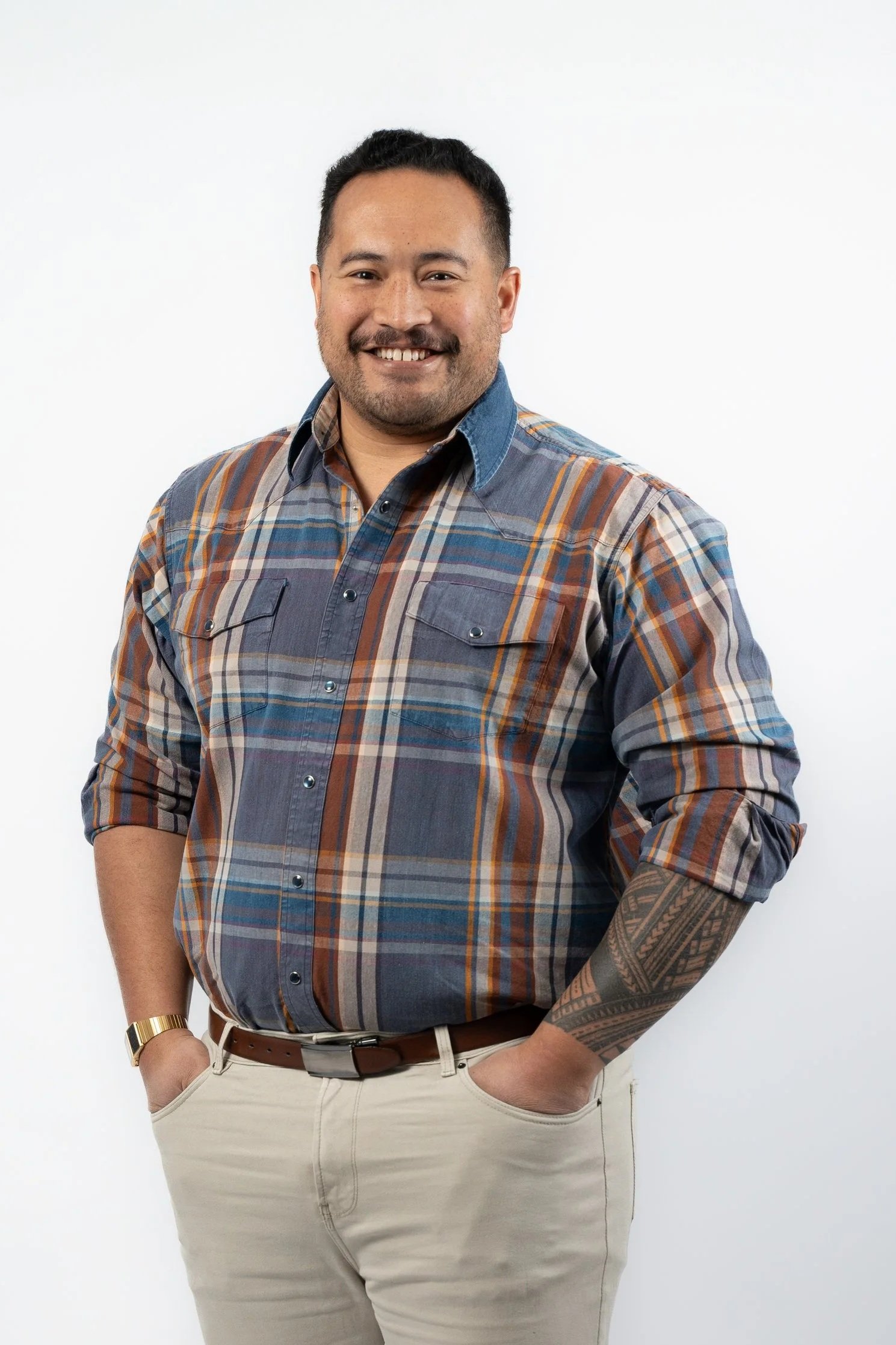 A smiling man with short black hair, a beard, and a tattoo sleeve on his right arm. He is wearing a plaid button-up shirt, beige pants, a brown belt, and a wristwatch, standing against a plain white background.