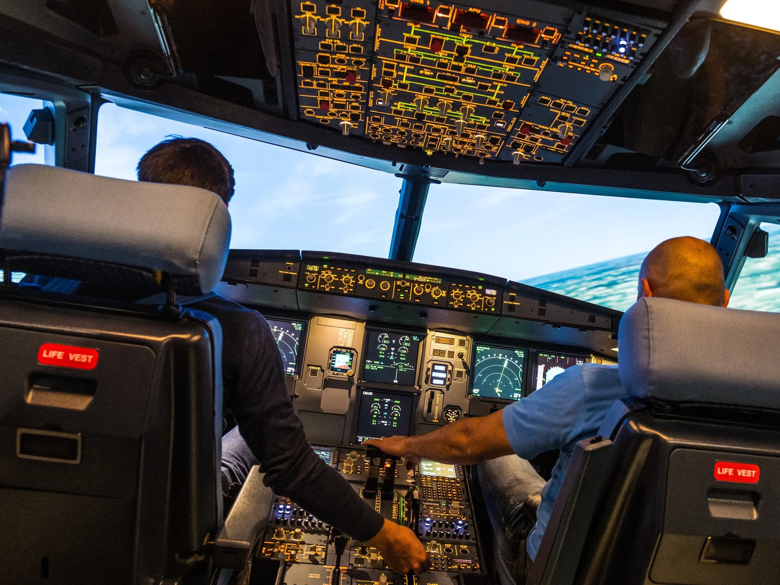 Inside the cockpit of an airplane with two pilots, numerous control panels, and screens showing flight data.