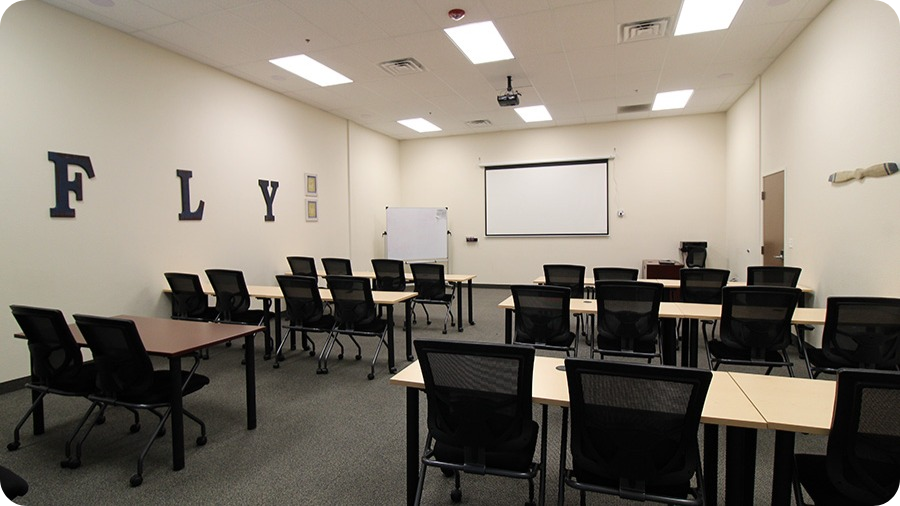 Empty classroom with black chairs and beige desks, whiteboard, projection screen, and framed letters spelling 'FLY' on the wall.