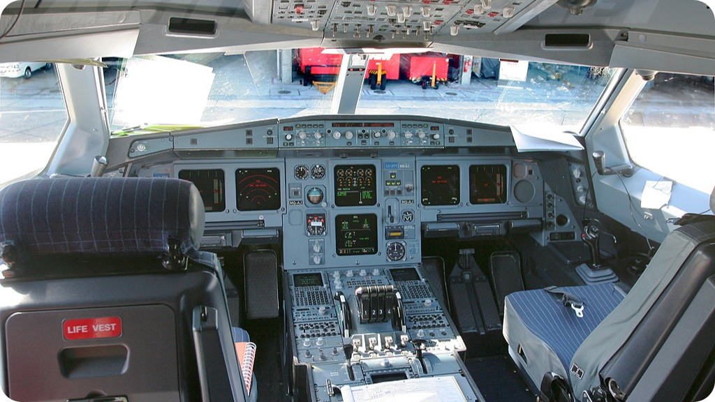 Inside view of an airplane cockpit with control panels, screens, and pilot seats, viewed from behind the pilots' seats at an airport gate.