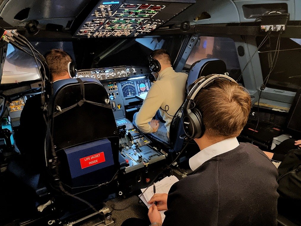 Three pilots inside an airplane cockpit with various controls and instruments, wearing headsets, with one pilot taking notes and others adjusting controls.