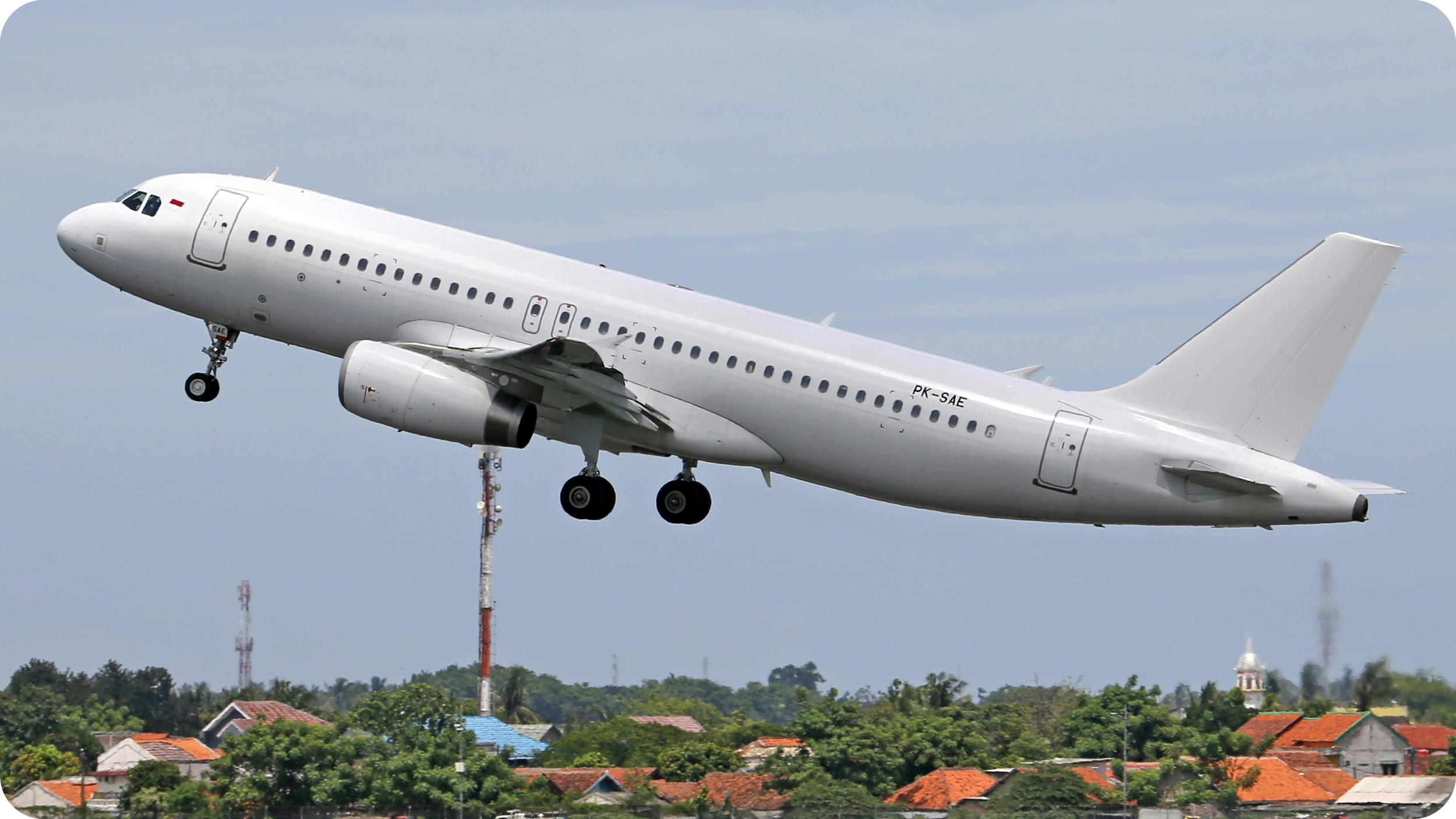 A commercial airplane in flight, with landing gear extended, flying over a residential area with houses and trees.
