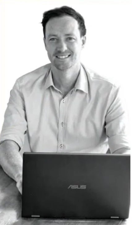 A smiling man with dark hair and a beard, wearing a light-colored shirt, sitting at a desk with an open ASUS laptop.