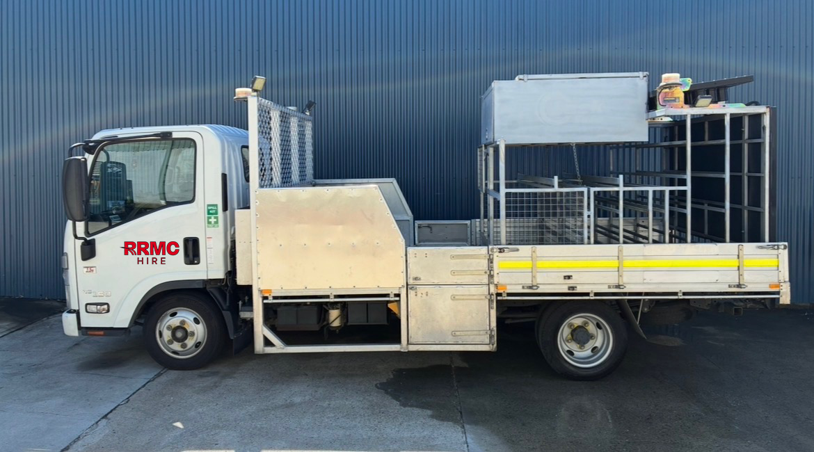 Work vehicle with metal racks and compartments, labeled 'RRMC HIRE', parked near a blue industrial building.