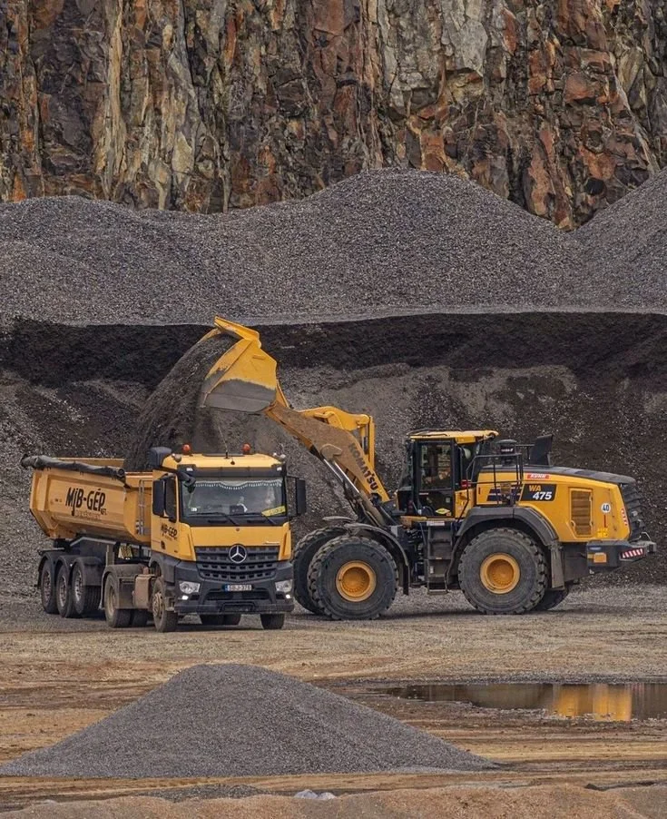 Construction site with a yellow wheel loader transferring gravel into a yellow dump truck, with large gravel piles and rocky cliff in the background.