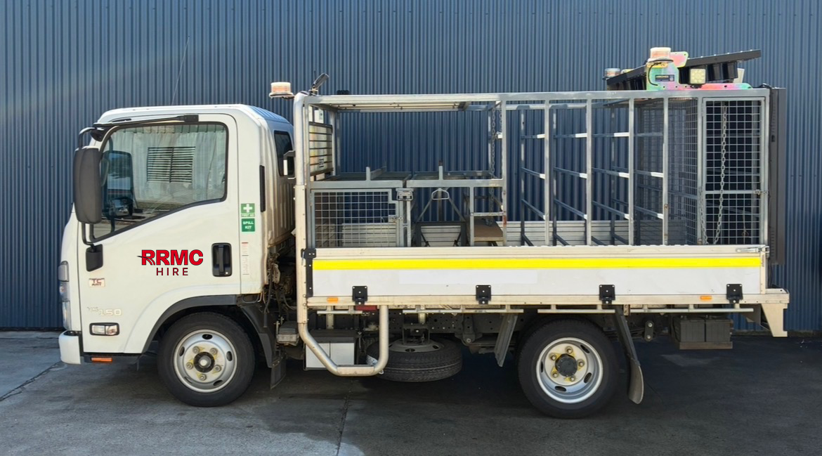 A white utility truck with cage and equipment on the back, parked in front of a blue corrugated metal wall.