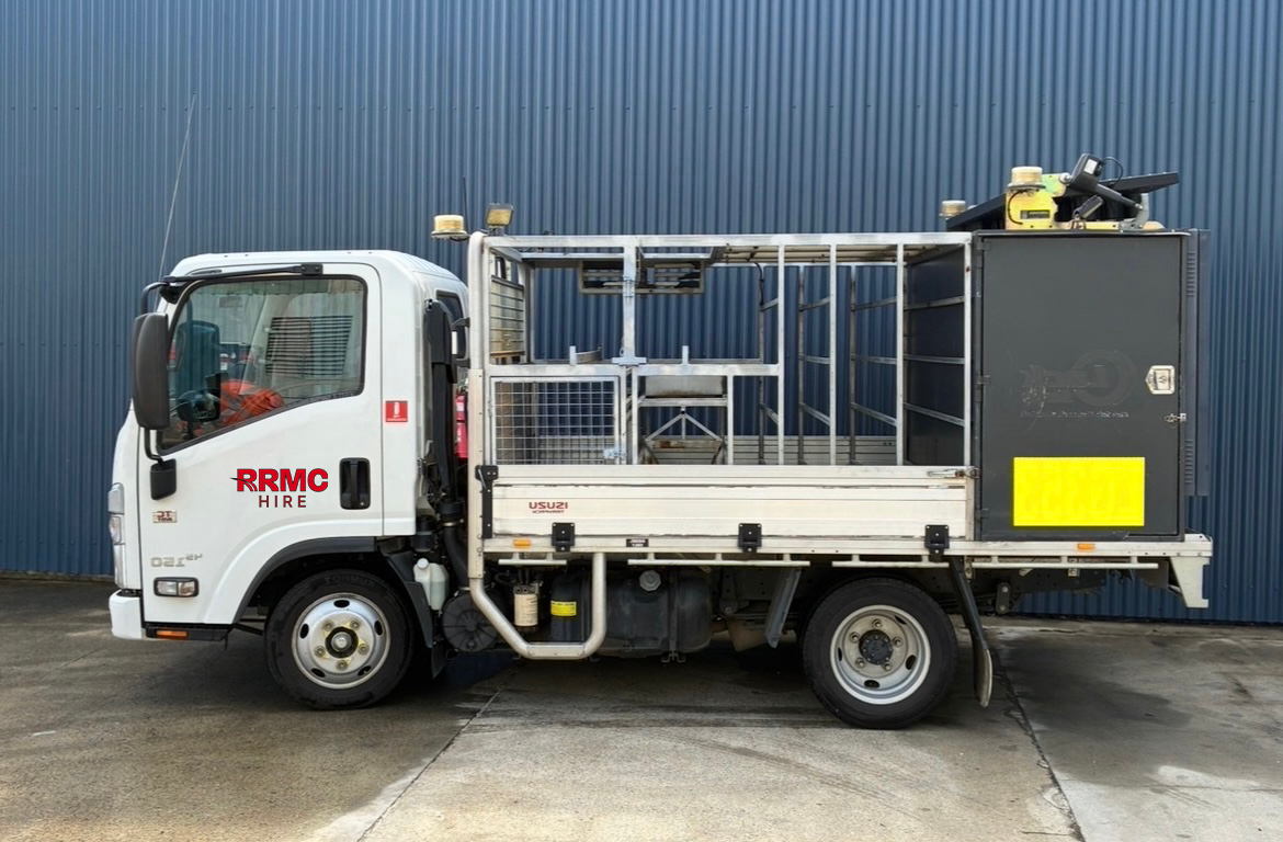 A white utility truck with empty cargo area parked on concrete ground in front of a blue corrugated metal wall.