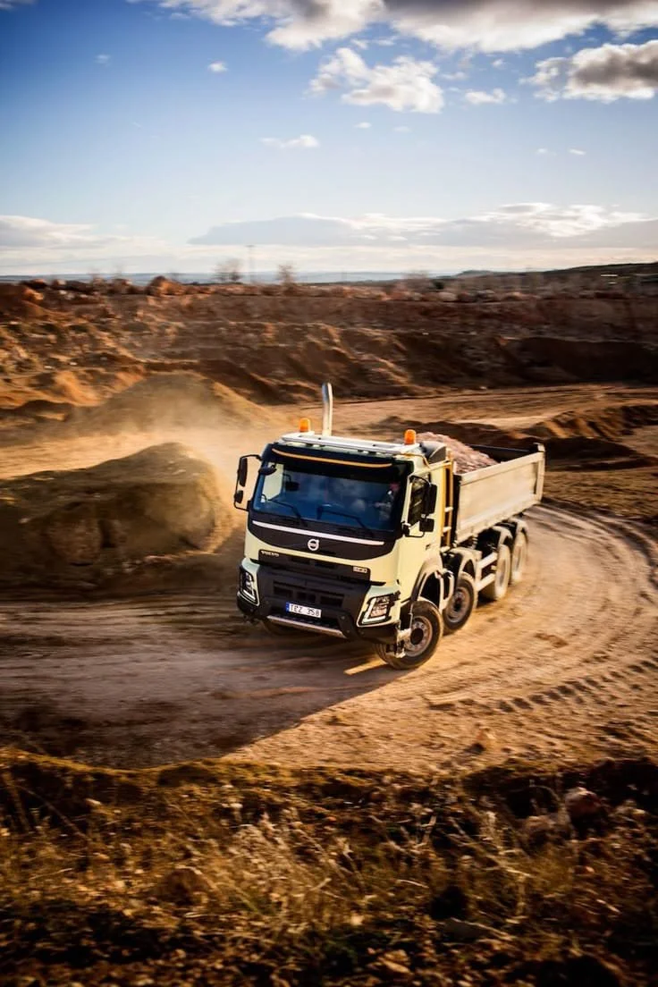 A white dump truck driving on a dirt road in a rugged, open landscape with hills and sparse vegetation.