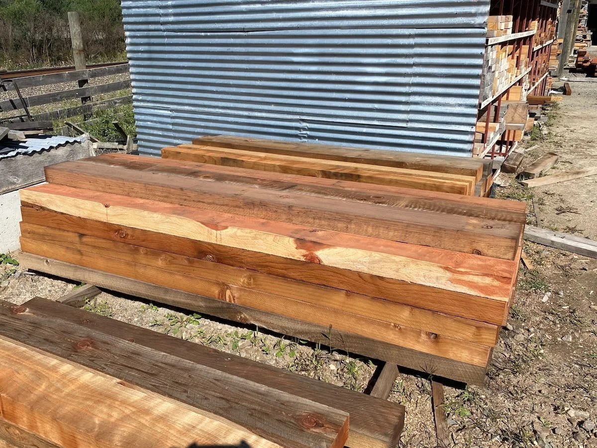 Stacks of cut wooden planks and beams outdoors in a lumber yard, with a corrugated metal wall in the background.