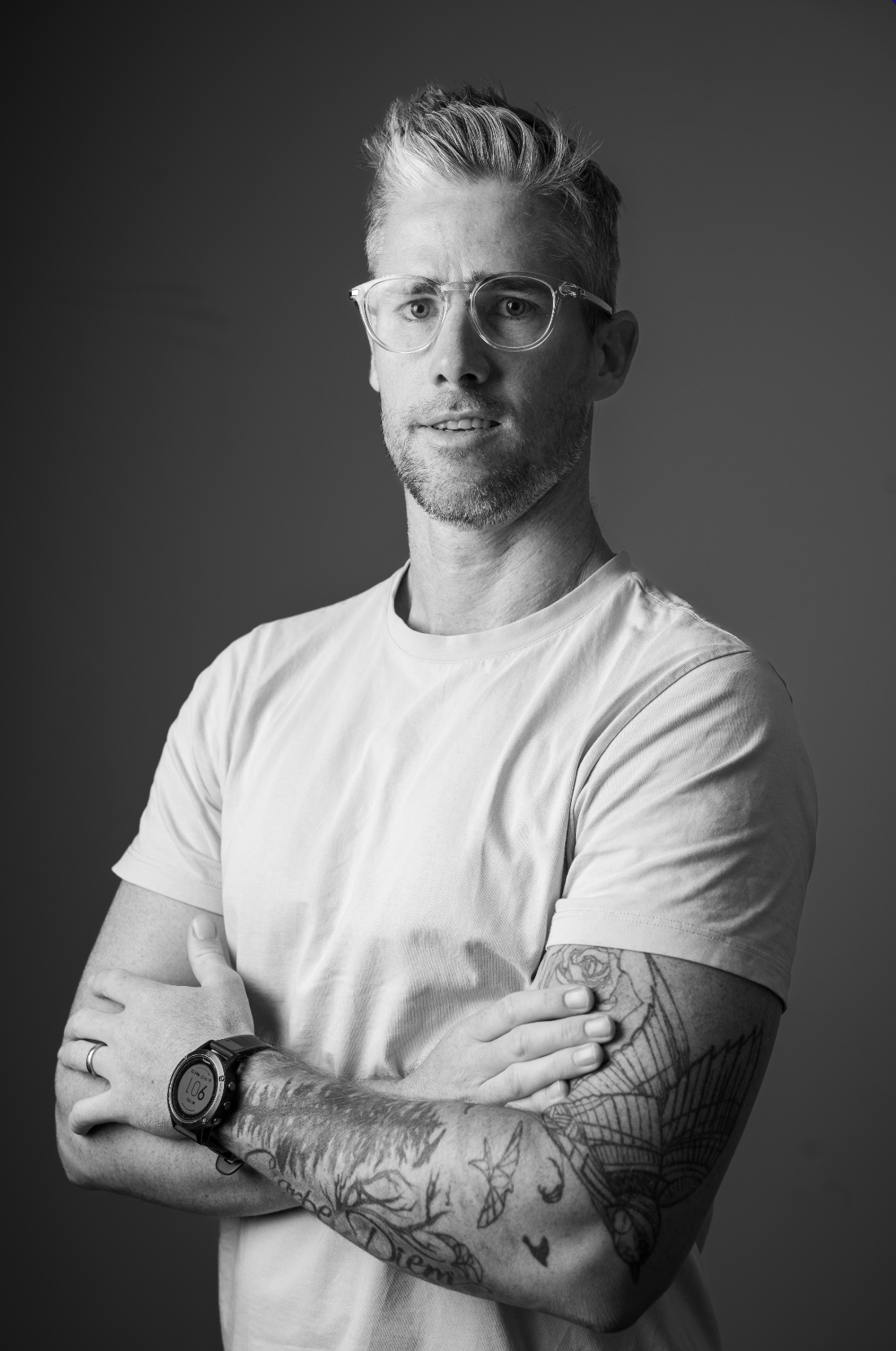 Black and white portrait of a man with glasses, short hair, and tattoos on both arms, wearing a plain t-shirt, standing with arms crossed against a plain background.