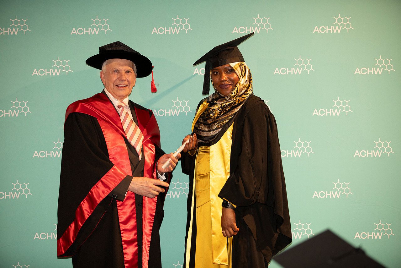 A graduate in a yellow gown and black cap receiving a diploma from a university official in academic regalia at a graduation ceremony.