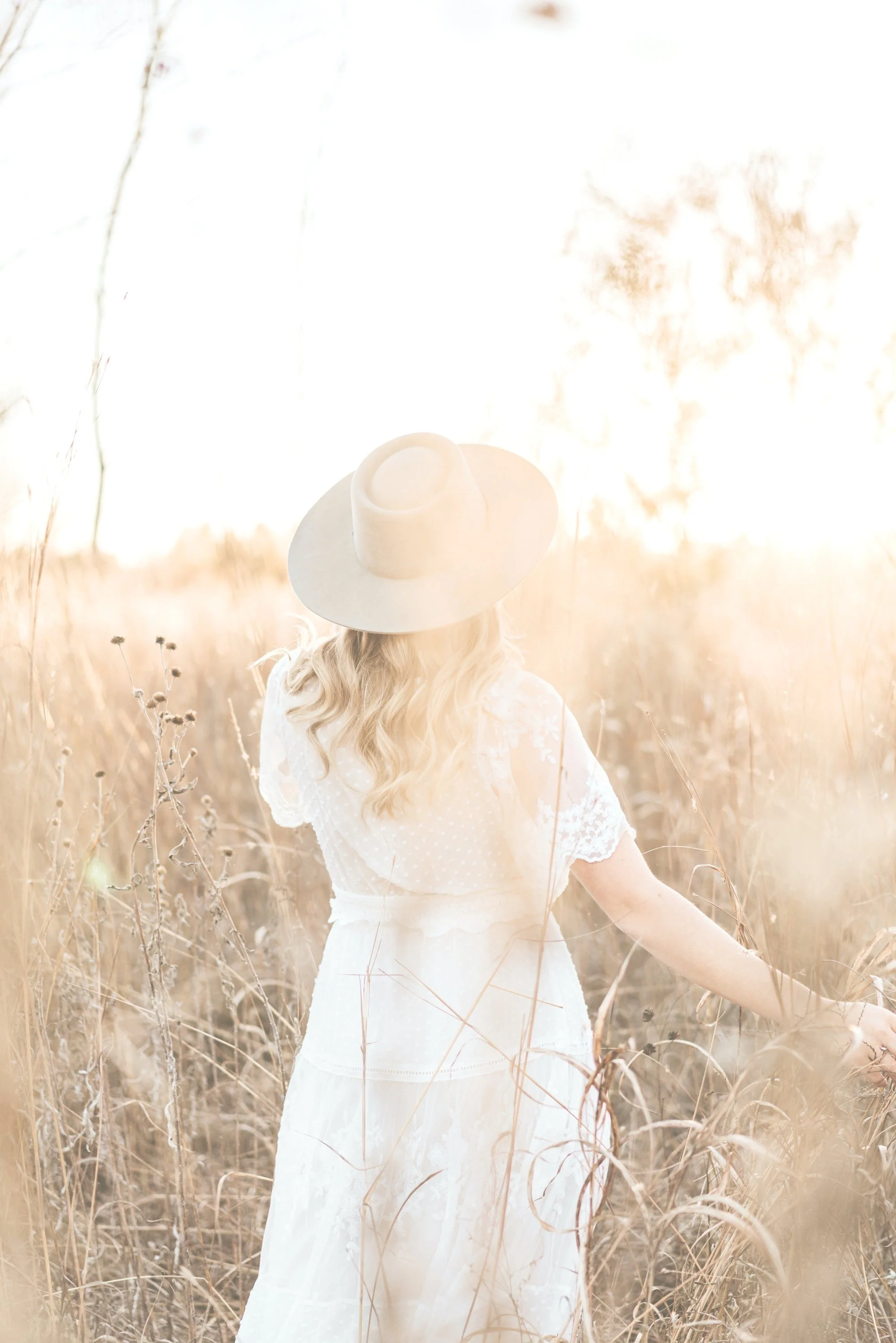 A woman with long, wavy blonde hair wearing a white dress and a wide-brimmed hat, standing in a sunlit field of tall grass, facing away from the camera.