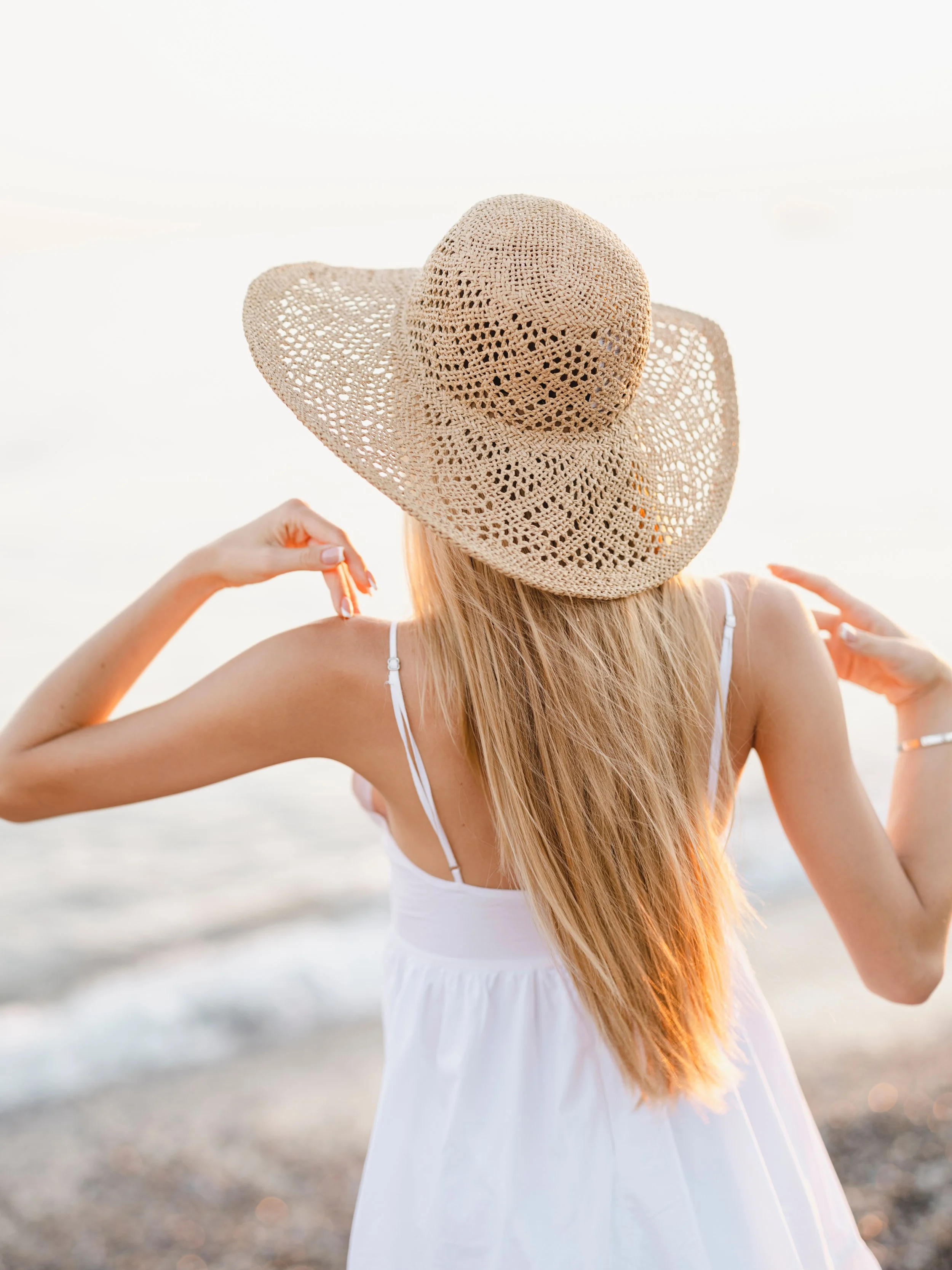 Woman with long blond hair wearing a white dress and a large woven sun hat, standing on a beach near the water, during sunset.