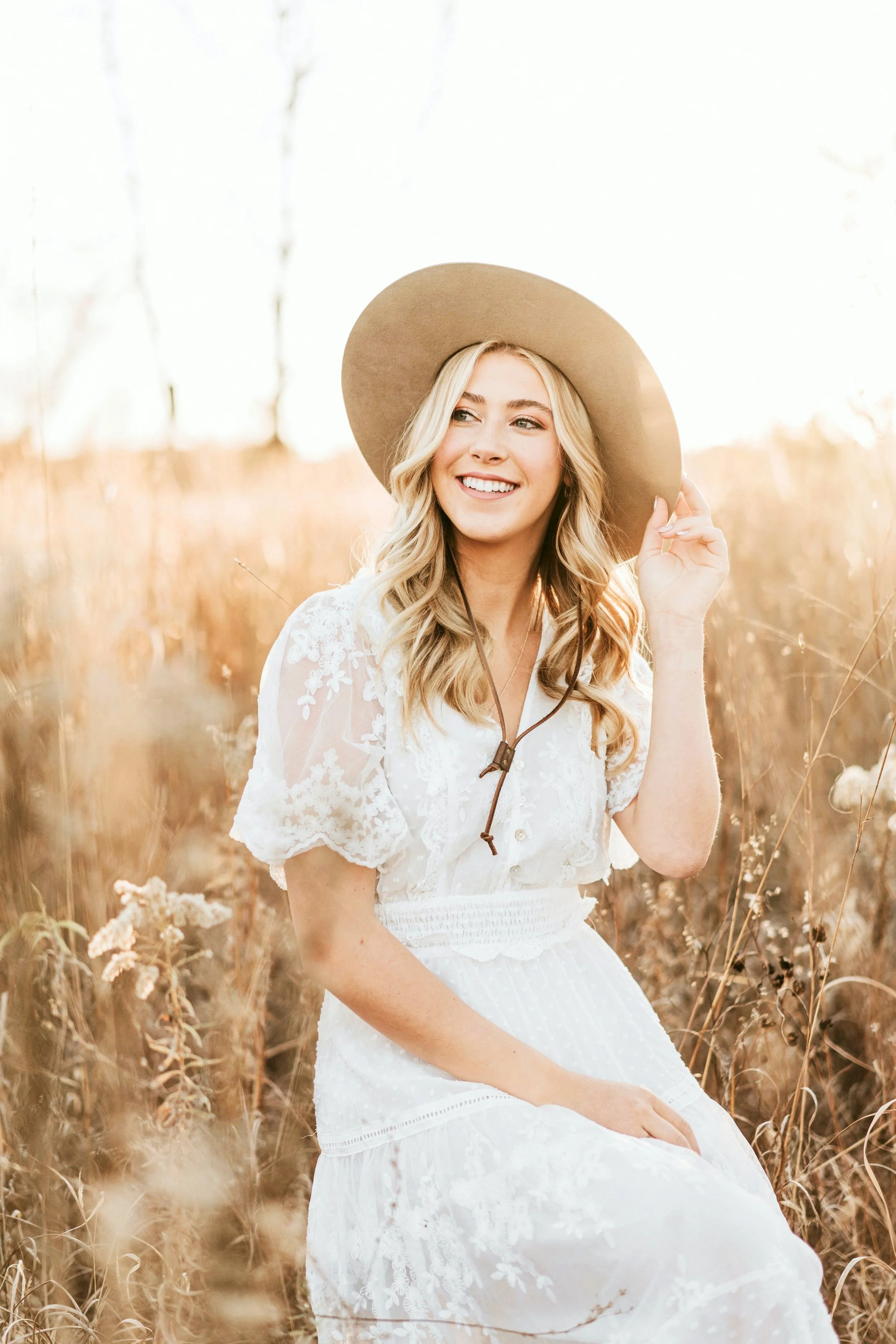 A young woman with blonde hair sitting in a field of tall, dry grass during sunset, wearing a white lace dress and a wide-brimmed tan hat, smiling and looking into the distance.