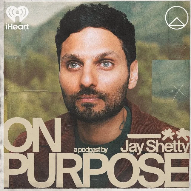 Close-up of a man's face with short dark hair, beard, and blue eyes, on a podcast cover titled "On Purpose" by Jay Shetty, with a green and brown blurred outdoor background.