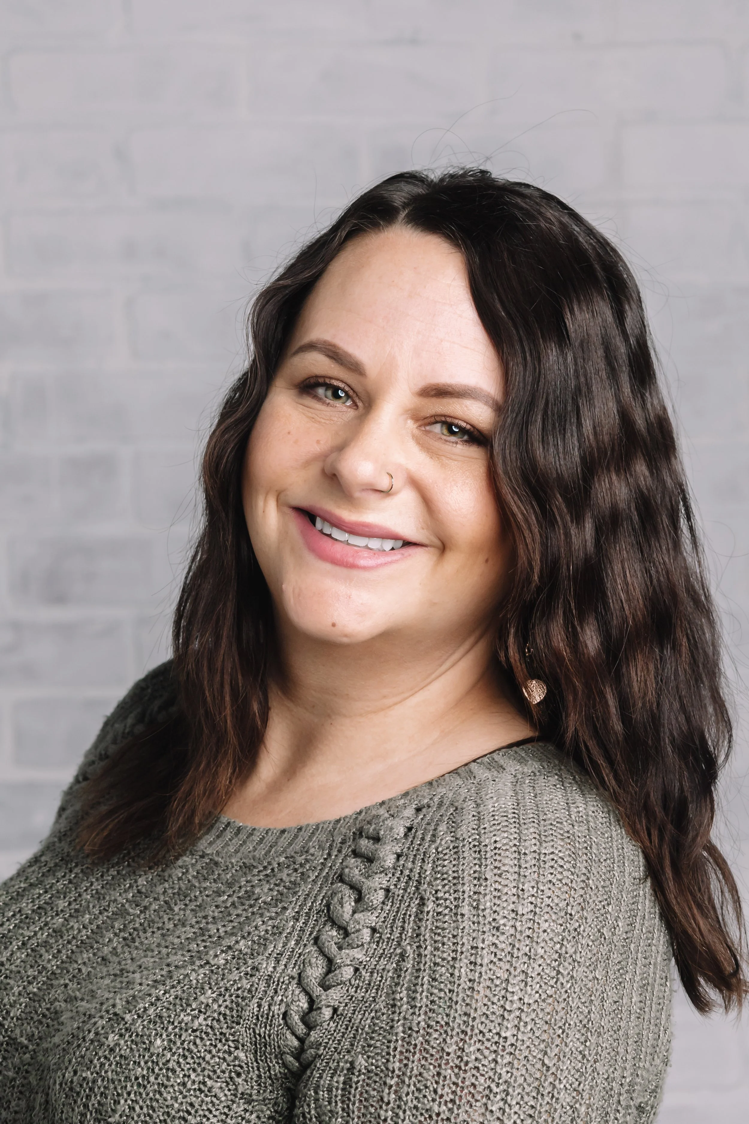 A woman with long dark wavy hair, light skin, and a nose piercing smiling at the camera, wearing a gray knitted sweater against a light gray brick wall.