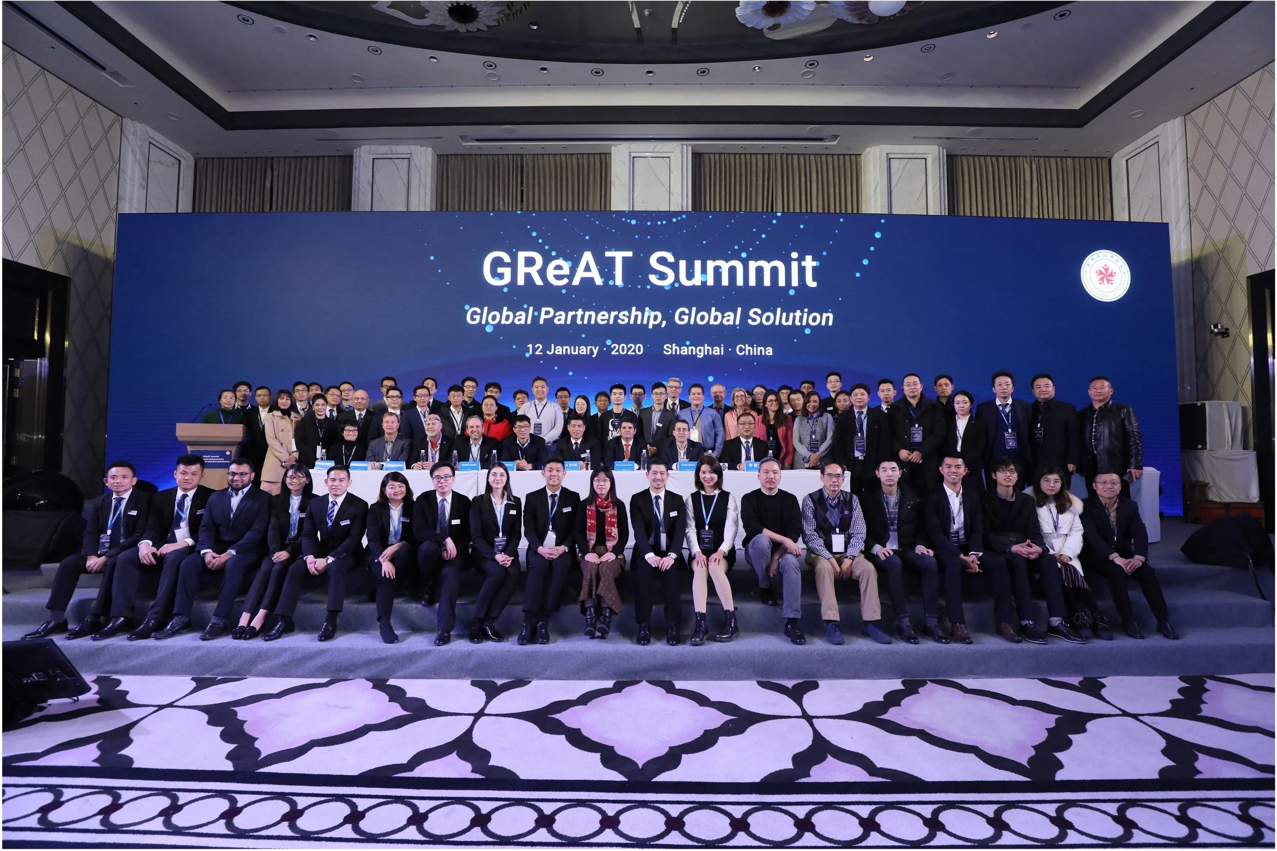 A large group of people is gathered on a stage for a photo at the GReAT Summit held in Shanghai, China, on January 12, 2020. They are posing in front of a large blue digital screen displaying the event name and details.