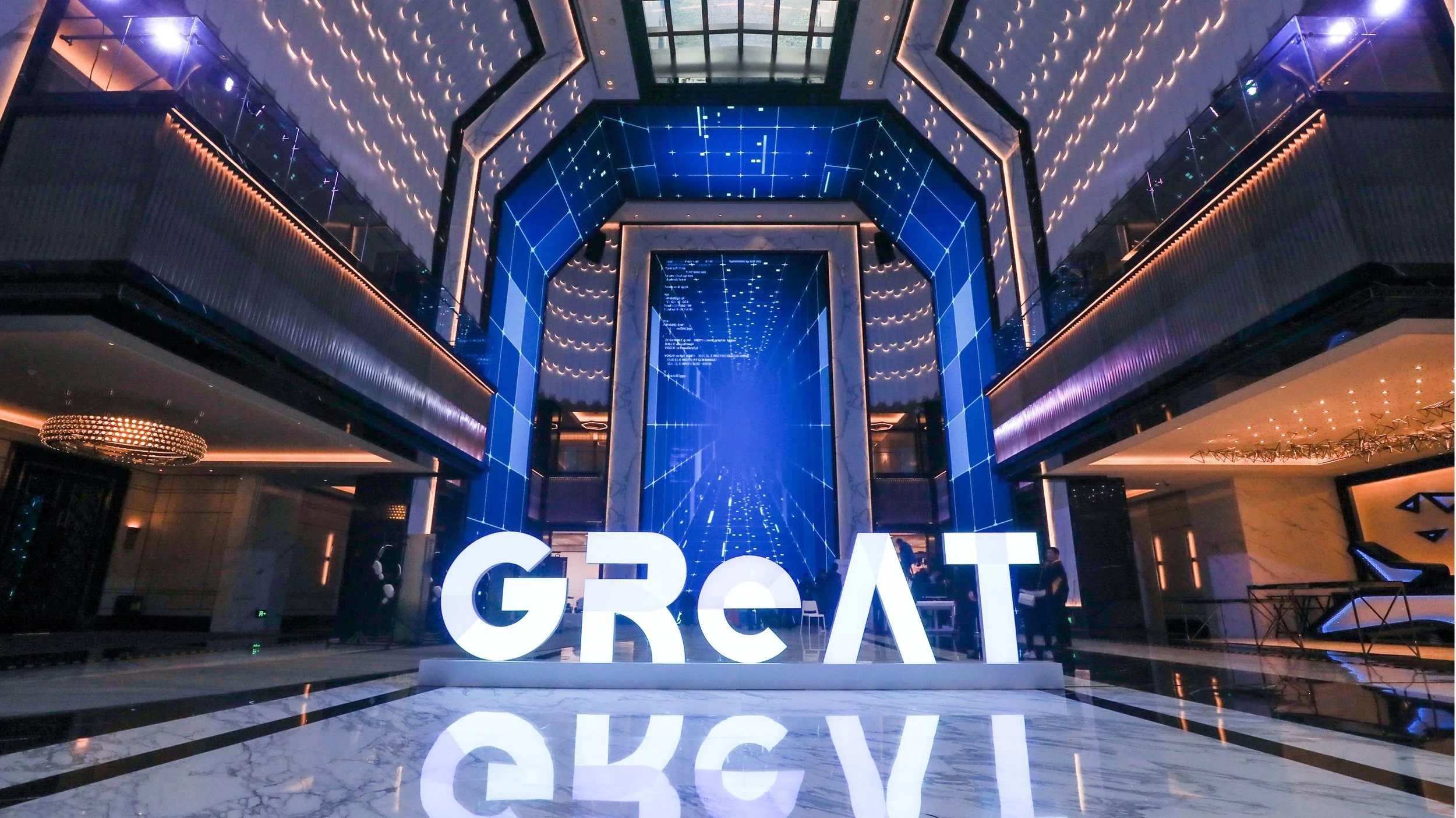 Indoor modern lobby with large illuminated sign spelling 'GREAT', glass and marble design, blue digital screen wall, and people in background.