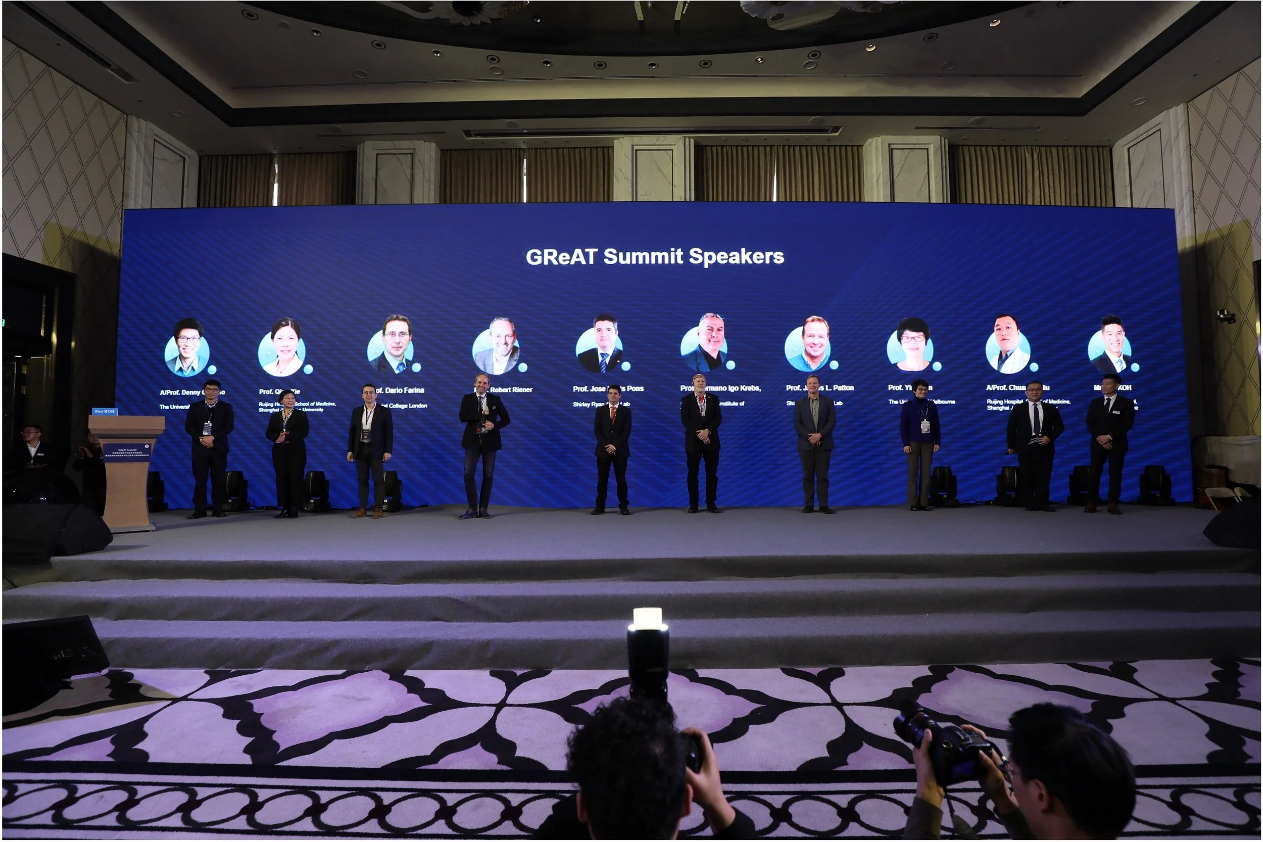 A group of people standing on a stage in front of a large screen displaying headshots and names of speakers at the GReAT Summit. The stage has a carpeted surface and the background features a high ceiling and elegant decor.