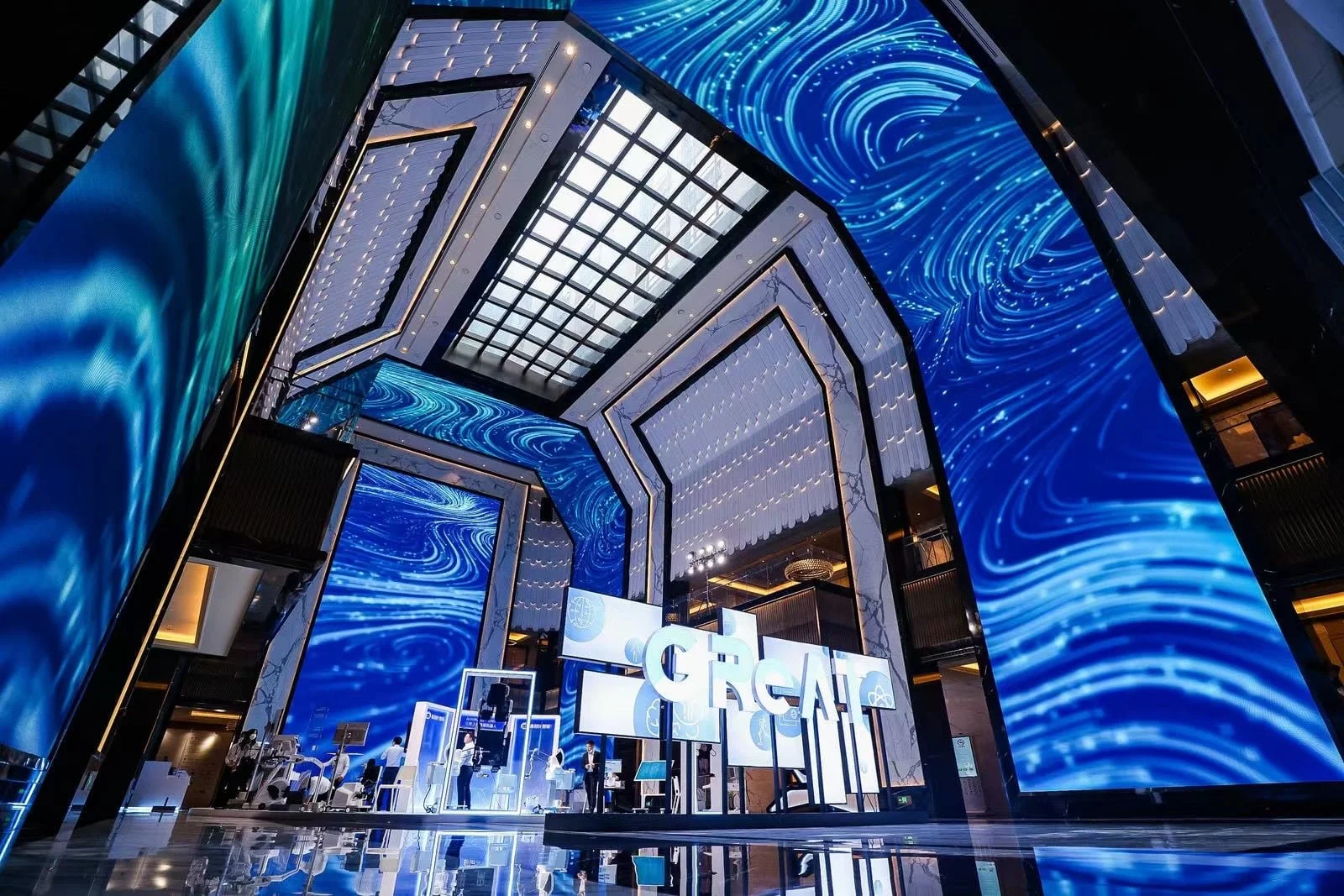 Inside a modern, upscale building with large digital screens displaying swirling blue patterns and the word 'CREATE' in big letters, featuring a high ceiling with geometric design and an exhibition or event setup with people and displays.