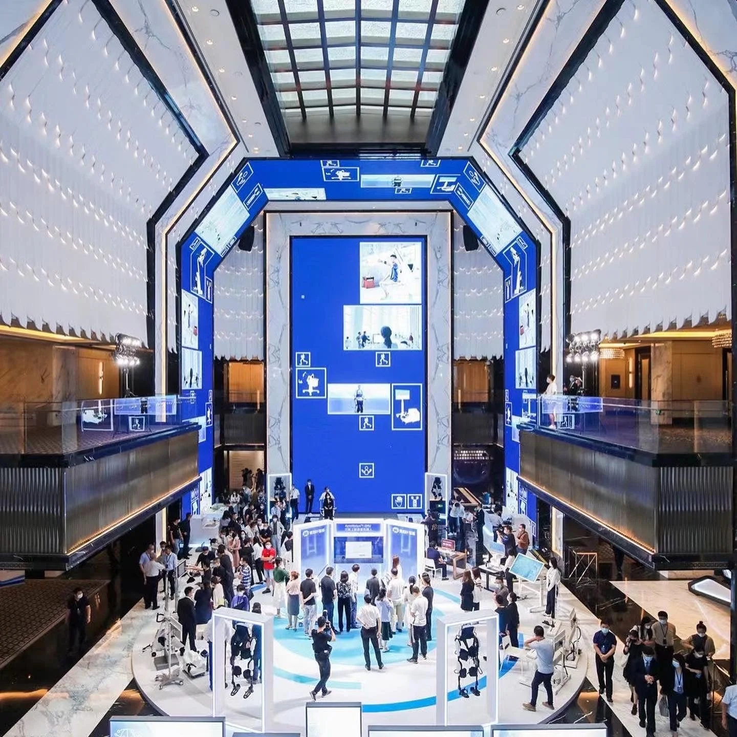Inside a modern building with a spacious atrium, blue and white signage, and many people attending an event or exhibition.