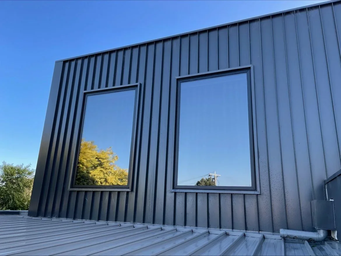 Close-up of a modern metal building facade with two rectangular windows reflecting trees and a clear blue sky.