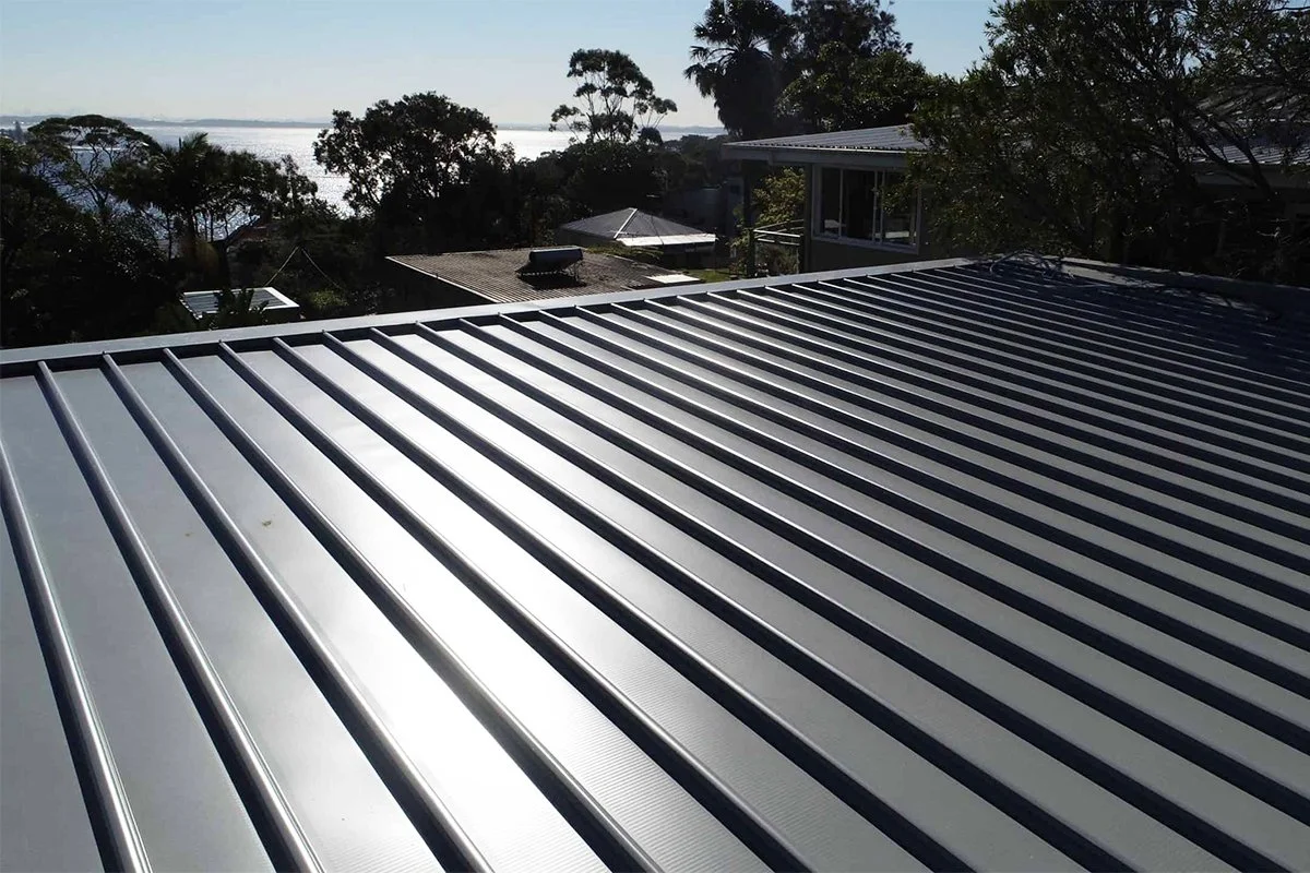 Silver metal roof with vertical ridges, surrounded by trees and with a view of a body of water in the distance.