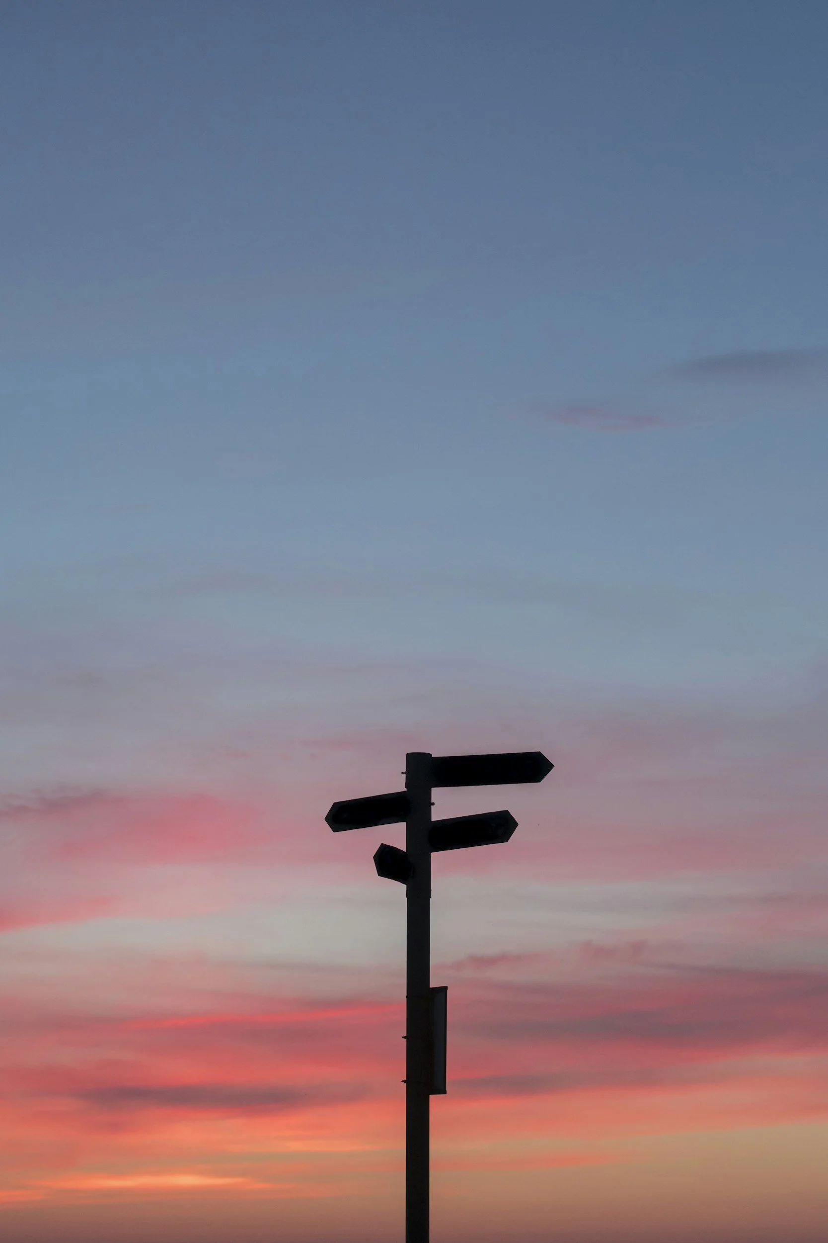 Silhouette of a signpost with directional arrows against a colorful sunset sky with pink, purple, and blue hues.