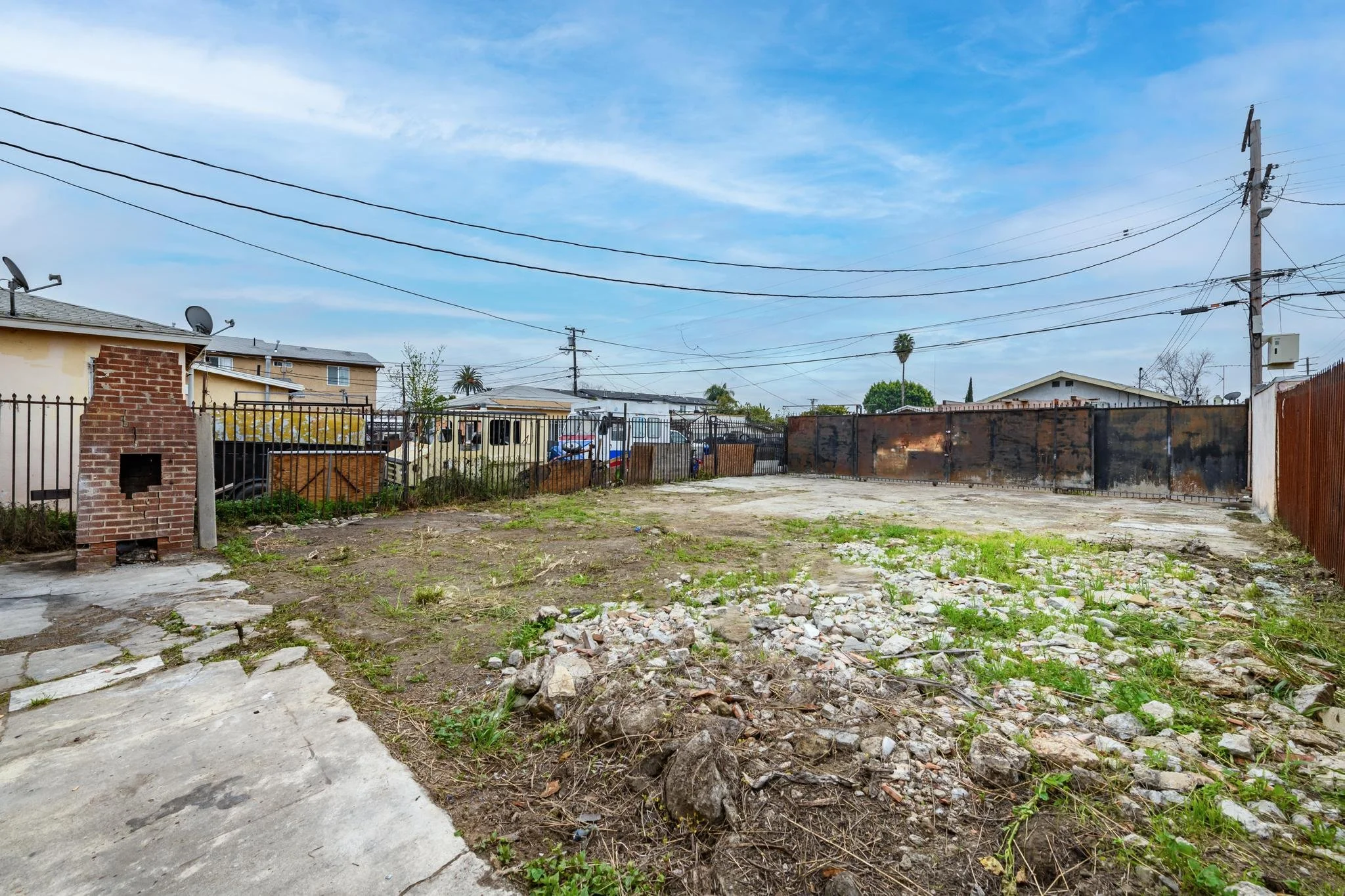 Empty backyard with dirt and sparse grass, surrounded by fences, with utility poles and houses in the background under a partly cloudy sky.