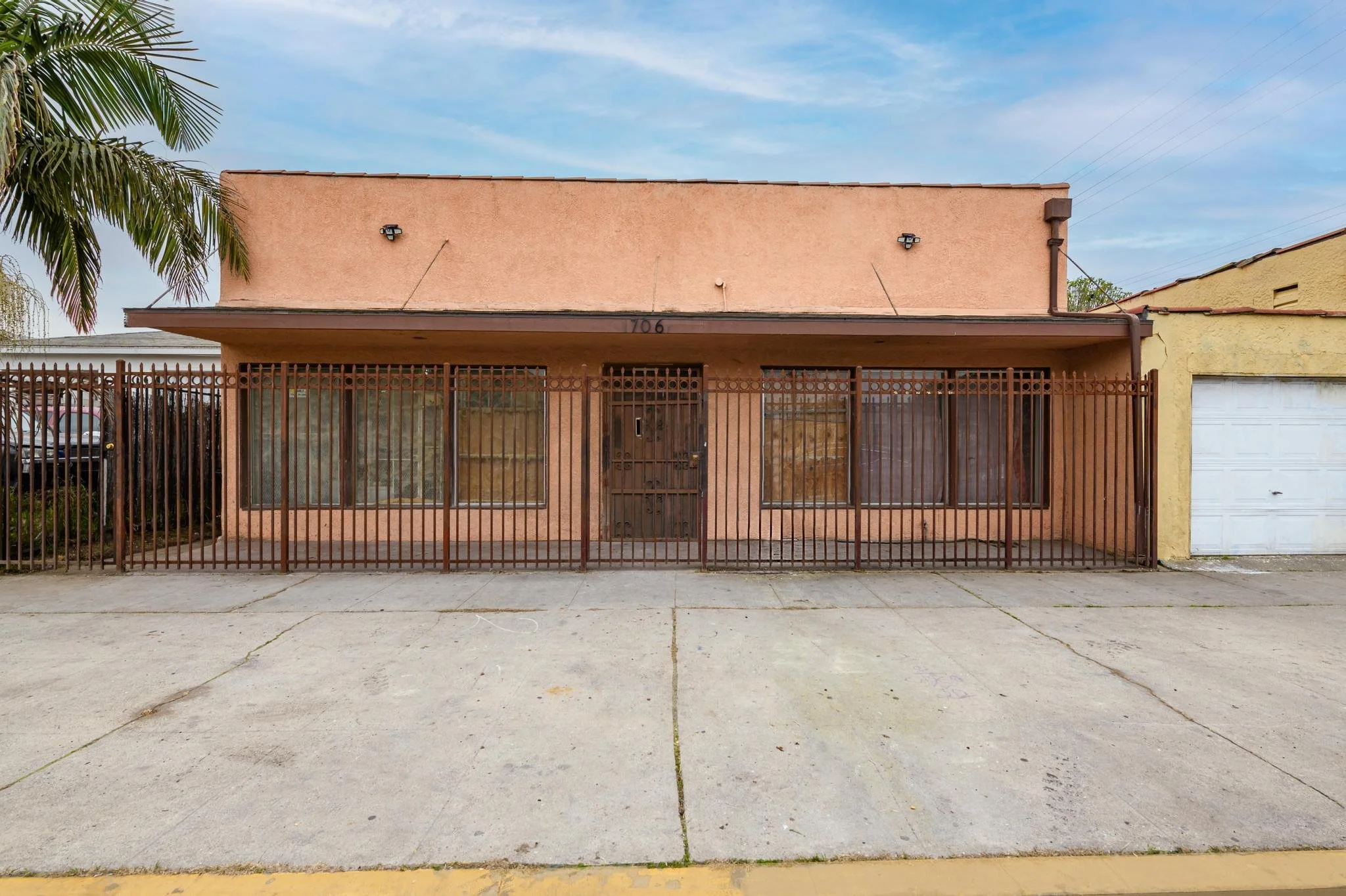 A pink single-story building with large windows, a brown door, and a metal fence in front. Adjacent to a yellow building with a garage door. A sidewalk and street are visible in the foreground.