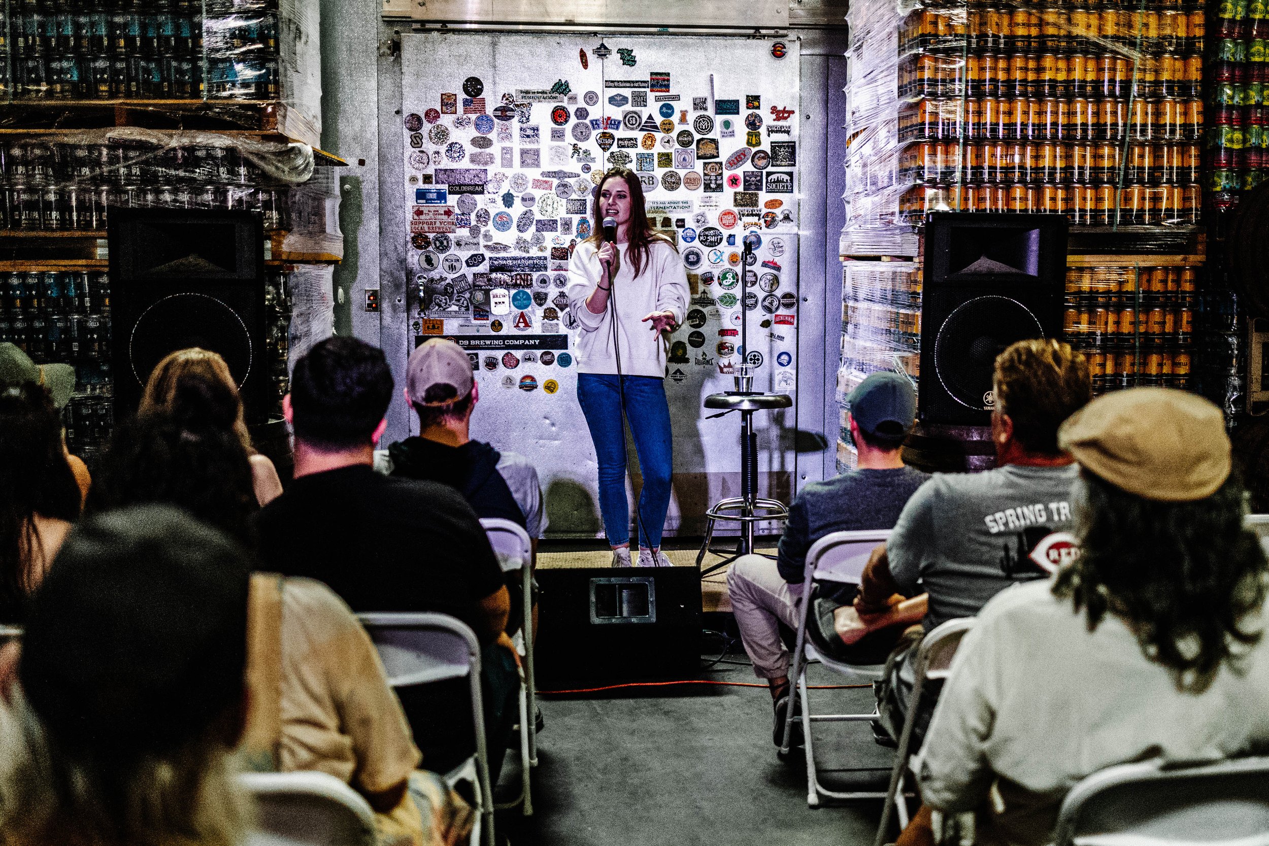 A woman performs stand-up comedy on stage in front of an audience in a brewery or bar setting, with large stacks of canned drinks and stickers on the wall behind her, while holding a microphone.