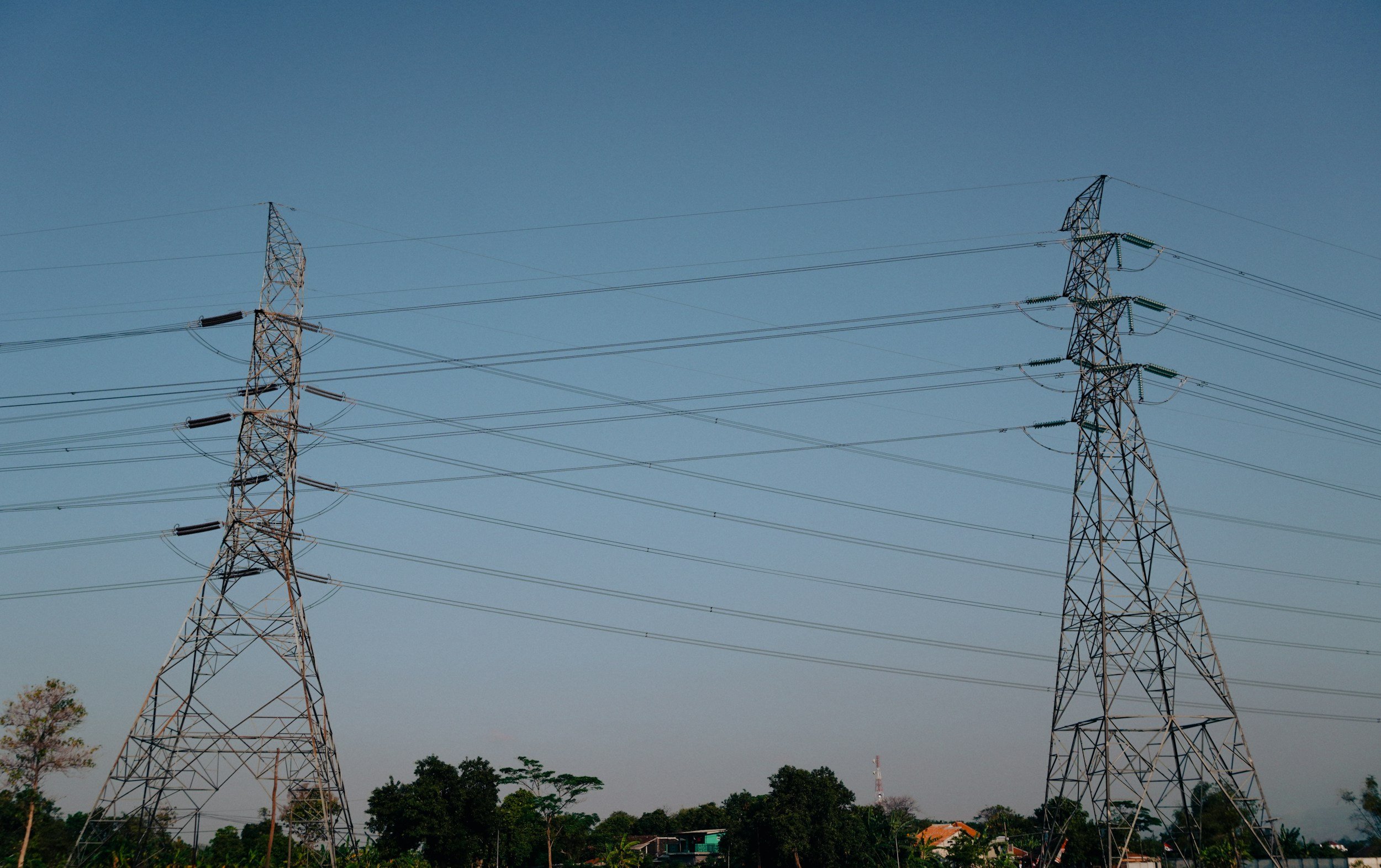 View of multiple electrical transmission towers against a clear blue sky with some greenery and houses in the lower background.