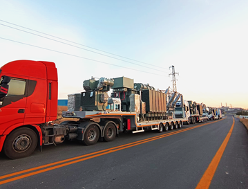A red semi-truck hauling military vehicles on a flatbed trailer along a two-lane highway at sunset.